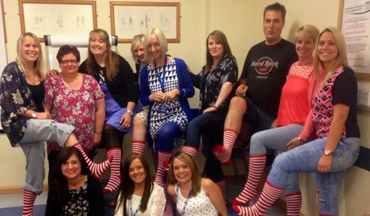 Thirteen smiling people, some sitting and some kneeling, pose with red and white striped socks pulled up to their knees, in an office-like setting.