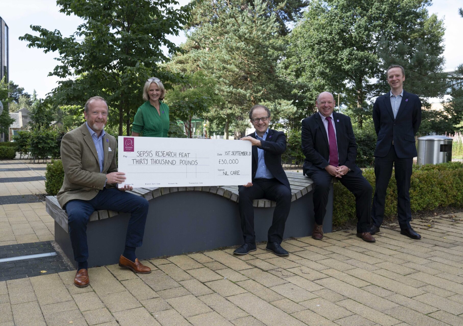 Five individuals present an oversized cheque for "SEPSIS RESEARCH FEAT THIRTY THOUSAND POUNDS Date 1ST SEPTEMBER 21 £30,000 Signed: NL CARE" on a paved area with trees and bushes.