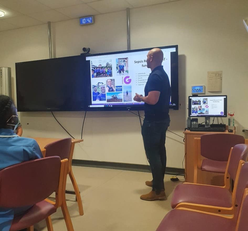 A bald man presents to a woman in a blue uniform, standing beside a large screen displaying "Sepsis R fun" and various images, in a room with empty chairs.