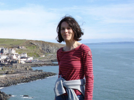 A woman stands smiling at the camera, with a coastal village and rocky coastline on a sunny day in the background.