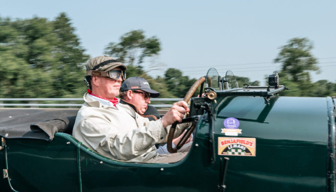 Two men are driving a dark green vintage car, blurred trees and a blue sky are in the background.