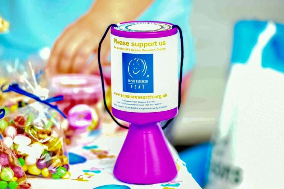 A purple and white collection tin for "Sepsis Research FEAT" sits on a patterned white surface surrounded by candy bags.