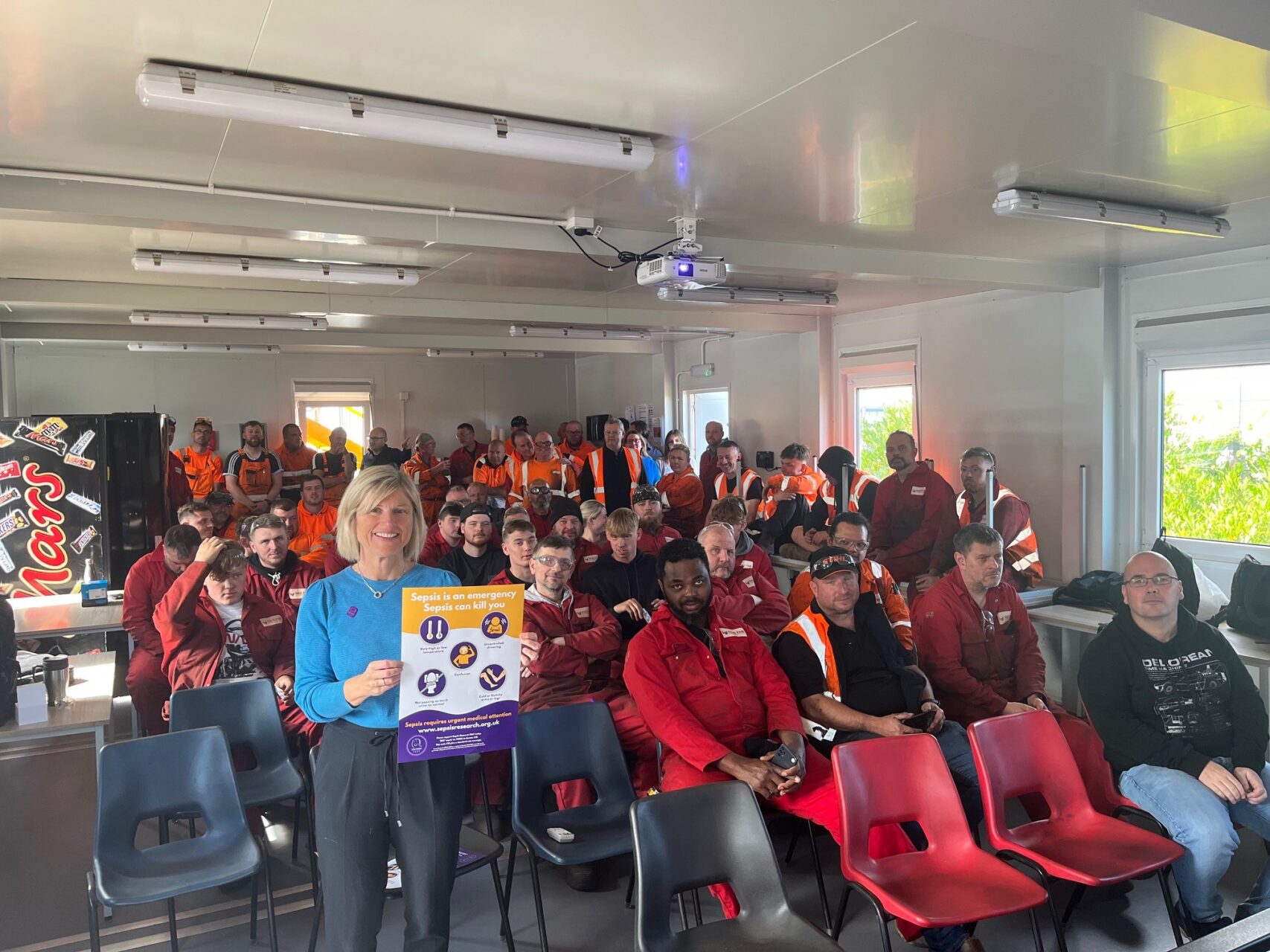 A woman in a blue top and dark pants holds a "Sepsis is an emergency, Sepsis can kill you" sign, addressing a group of men in red and orange overalls seated in a bright room.